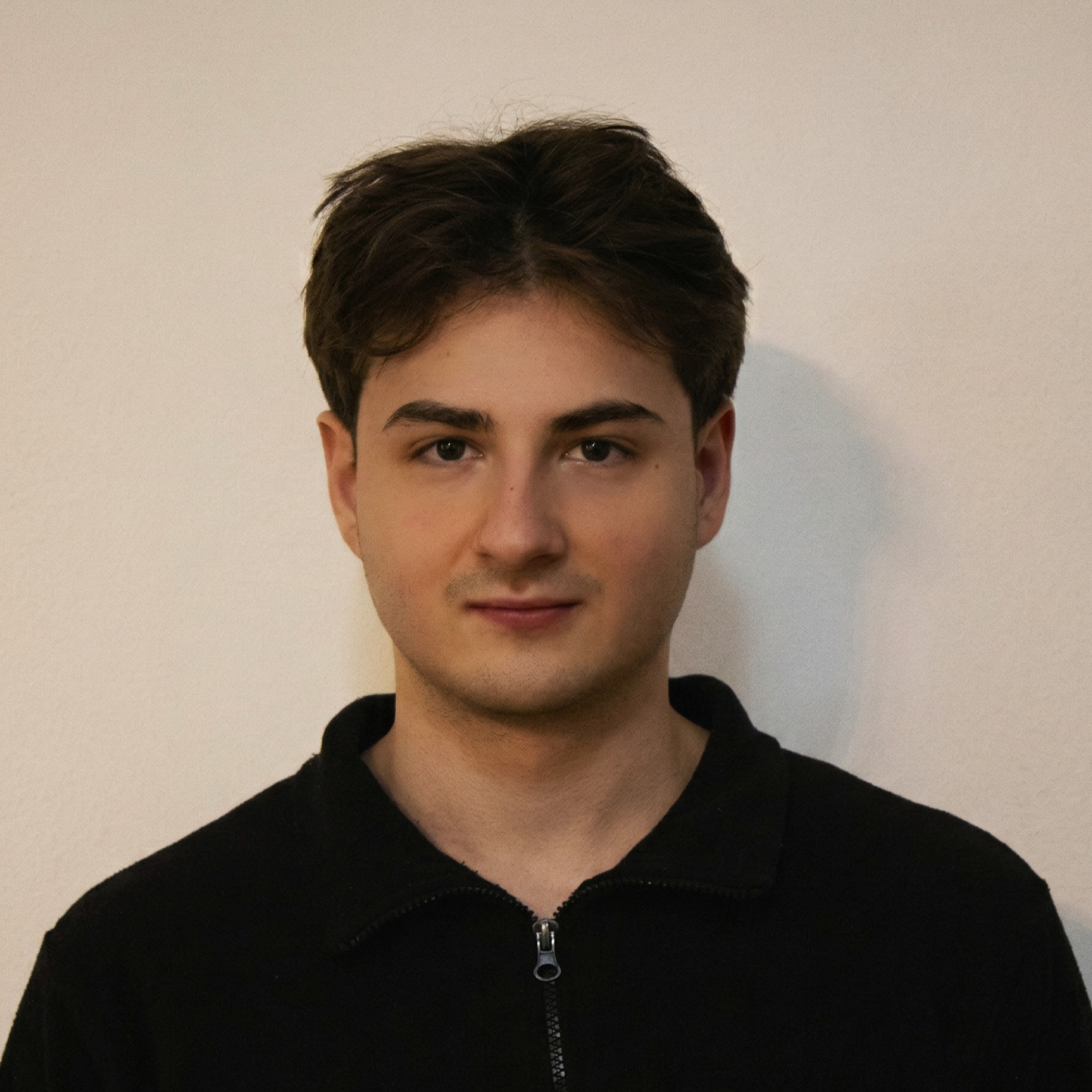 Portrait of a young man with wavy brown hair wearing a black polo shirt, standing in front of lush green foliage. Professional and confident outdoor headshot.
